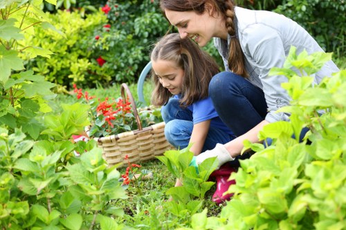 A woman and a young girl are gardening together in a lush, well-maintained outdoor space, with various green plants, bushes, and flowering shrubs surrounding them. The woman is crouched down, smiling, and holding a small gardening tool, while the girl, also crouching and engaged, is collecting red flowers into a wicker basket. In the background, there is a neatly edged flower bed with vibrant red blossoms and dense green foliage, alongside a paved pathway with a textured surface. The garden features a mixture of natural soil areas and manicured lawn sections, with sunlight filtering through the leaves, creating a bright and welcoming outdoor environment. This scene exemplifies typical gardening activities in a suburban garden setting, aligned with landscaping and lawn care services in Marylebone or similar areas in London.