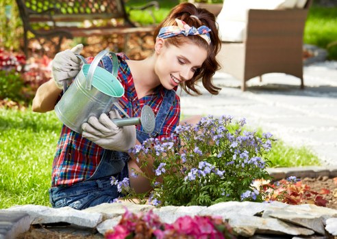 A young woman with brown hair tied back and wearing a colorful plaid shirt, grey gardening gloves, and a light headband, is working in a well-maintained front garden in Marylebone. She is watering a cluster of small, light purple flowering plants with a silver watering can, smiling as she tends to the garden bed. The garden features a neatly edged flower bed with various plants and flowers, bordered by smooth, grey stone edging. In the background, there is a patio area with outdoor furniture, including a wooden chair, and lush green grass surrounding the paved area. The scene is set on a bright, sunny day, with natural light illuminating the garden and creating shadows from nearby trees or plants. The environment appears tidy and cared for, reflecting professional gardening standards, as seen through the clean paving and healthy plants, illustrating typical garden maintenance tasks in a residential outdoor space within the Marylebone neighbourhood. This image aligns with landscaping and gardening services offered by Landscapers Marylebone, featuring outdoor plant care and garden upkeep in London’s Marylebone area.