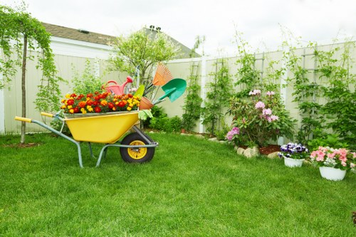 A well-maintained backyard garden with a lush, green lawn bordered by flower beds and potted plants, including pink and purple blooming flowers. A bright yellow wheelbarrow filled with colorful gardening tools and flower pots sits on the grass, indicating ongoing gardening work. Behind the garden, a white panel fence surrounds the space, with climbing plants and shrubs adding greenery along its length. In the background, a small tree and a hedge line further enclose the garden, which is illuminated by natural daylight under a partly cloudy sky. The environment appears neat and organized, reflecting professional gardening and outdoor maintenance standards typical of services provided by Landscapers Marylebone, supporting local sustainability and recycling efforts within a residential area in London.