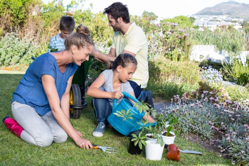 A family working together in a landscaped garden, with a woman planting a small shrub in a neatly maintained grass area, children assisting nearby with gardening tools and a watering can, while a man in the background observes. The garden features a variety of flowering plants, lavender, and small bushes within a bordered flower bed, with a paved patio area visible on one side. The lush lawn is well-kept, bordered by natural pathways and low hedges, with trees and distant buildings suggesting a residential outdoor space in Marylebone. The scene is well-lit with natural daylight, indicating a clear, mild weather day, typical of spring or summer, emphasizing outdoor gardening and maintenance activities conducted by Landscapers Marylebone.
