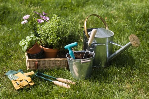A neatly maintained front garden in Marylebone featuring a lush green lawn with dense, evenly cut grass. In the foreground, there is a small flower bed bordered by a low edge, containing potted plants and flowering shrubs with purple and green foliage. To the right, gardening tools such as pruning shears, gloves, and small hand trowels are arranged on the grass, alongside a metal watering can with a wooden handle, indicating recent outdoor maintenance activities. The background includes a well-kept hedge line separating the garden from neighboring properties, and mature trees with full green leaves provide shade. The scene is captured on a bright day with natural sunlight illuminating the varied textures of the grass, foliage, and garden accessories, reflecting professional outdoor care typical of landscaped residential spaces in central London, near postcode W1H. The presence of gardening tools and watering equipment subtly supports the services offered by Landscapers Marylebone in professional garden maintenance and landscaping. The overall environment conveys a neat, inviting outdoor area suitable for relaxing or outdoor activities.