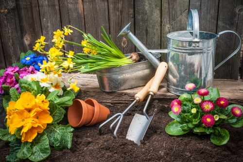 A garden scene featuring a freshly prepared flower bed with dark, rich soil, surrounded by colorful flowering plants including pink, yellow, and blue blossoms. In the background, there is a weathered wooden fence enclosing the outdoor space. Various gardening tools are arranged on the soil, including a metal watering can, a pair of hand trowels with light-colored handles, and a small hand rake. Two empty terracotta pots are stacked on the soil, and a metal container holds a bunch of yellow and white daffodils. The overall setting suggests ongoing gardening work, with a focus on planting and garden maintenance in a backyard or front garden area, typical of a residential garden in Marylebone, London, with natural lighting indicating a partly cloudy day.
