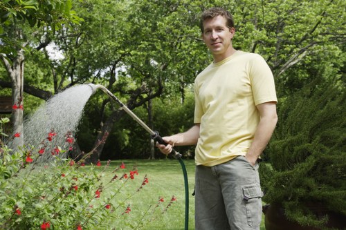 A man with light brown hair, wearing a pale yellow T-shirt and grey cargo shorts, stands in a lush, well-maintained garden surrounded by green trees and shrubs. He is holding a garden hose with a spray nozzle, watering a flower bed that contains small red flowers and various green plants. The garden features a neatly mowed grass lawn, a paved pathway visible to the right, and mature trees providing partial shade. The background includes dense foliage and a vibrant natural outdoor environment, suggesting a peaceful and tidy private garden space typical of a residential area in Marylebone. The scene is well-lit with natural daylight, indicating a clear weather day, and exemplifies routine outdoor maintenance that gardening services, such as those offered by Landscapers Marylebone, can assist with to promote sustainable and well-kept outdoor spaces.