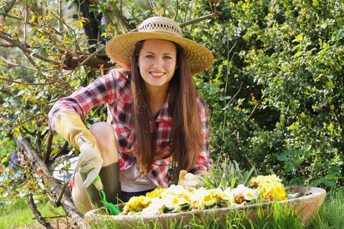 A young woman with long brown hair, wearing a wide-brimmed straw hat, a red and white checkered shirt, and gardening gloves, is kneeling in a well-maintained garden. She is tending to a flower bed containing a variety of blooming yellow, white, and purple flowers. The garden features a lush green lawn area in the foreground, edged by a curved flower bed with rich, dark soil and planted flowers. Behind her, there are small trees and leafy shrubs, providing a natural backdrop of foliage. The scene appears to be outdoors in bright, sunny weather which enhances the vibrant colours of the flowers and the greenery. The overall setting reflects a carefully landscaped garden space, typical of residential gardens in Marylebone, with an emphasis on flower bed creation and outdoor plant care, aligning with professional gardening and landscaping services offered by Landscapers Marylebone.