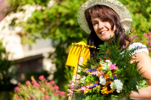 A smiling woman wearing a wide-brimmed straw hat stands in a lush garden, holding a vibrant bouquet of mixed flowers and greenery. Behind her, the garden features well-maintained grass with a dense, green hedge or shrubbery along the background, suggesting a private outdoor space in London, possibly in Marylebone. The environment appears bright and sunny, with natural light illuminating the scene and casting soft shadows. The garden has a mixture of natural textures, including a neatly trimmed lawn, flowering plants in the background, and garden tools such as a yellow rake visible near her. The overall setting reflects a thoughtfully designed outdoor area suitable for gardening and landscaping, aligning with services offered by Landscapers Marylebone, serving the local postcode area, enhancing exterior aesthetics and outdoor usability.
