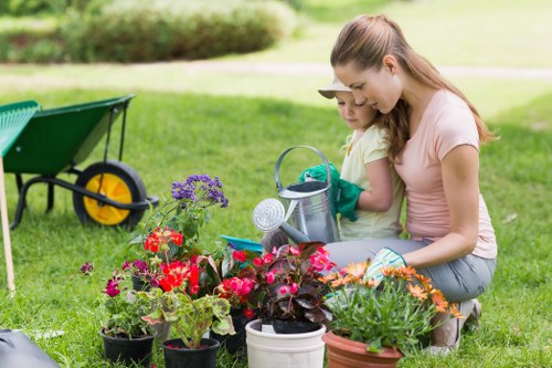 In a well-maintained garden in Marylebone, two young girls are engaged in gardening activities amidst a lush green lawn, with a background of soft-focus trees and shrubbery. The foreground features an assortment of potted plants and flowers, including vibrant red, pink, purple, and orange blossoms, set on the grass. One girl, wearing a light yellow t-shirt, holds a small watering can and gently waters the plants, while the other, dressed in a peach top, assists by holding the watering can. To the side, a green wheelbarrow with yellow handles is partially visible, resting on the grass, suggesting ongoing outdoor work. The garden layout includes a smooth lawn surface, with garden beds or borders not immediately visible but implied by the arrangement of flowers and potted plants. The weather appears mild and sunny, providing natural light that highlights the rich colours of the plants and enhances the outdoor environment's inviting atmosphere. This scene illustrates a typical garden setup suitable for gardening and lawn care services offered by Landscapers Marylebone, reflecting a healthy, landscaped outdoor space in central London postcode area, perfect for family gardening activities and outdoor maintenance.