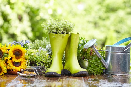 The image displays a garden scene with a weathered wooden outdoor table holding gardening tools and decorative elements. Prominently, there are a pair of bright green rubber gardening boots filled with small white and green flowers, positioned upright in the centre. To the left of the boots, a bunch of vibrant yellow and brown sunflower blooms rests on the table, adding a splash of colour. On the right side of the boots, there is a metal watering can with a long spout and a handle, partially visible, suggesting outdoor watering or garden maintenance activity. The background features lush greenery, blurred to emphasize the foreground objects, indicating an outdoor garden environment with soft, natural sunlight filtering through. The scene conveys a well-maintained garden space, with elements typical of gardening services in Marylebone, combining practical tools and decorative touches, set against a backdrop of vibrant, healthy foliage suitable for lawn and flowerbed care, typical of professional landscaping and gardening in London neighborhoods.