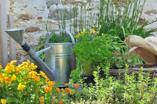 The image depicts a small, well-maintained garden area with a rustic stone wall in the background. In the foreground, vibrant yellow and orange flowering plants are visible, adding bright natural tones to the scene. Behind these, there is a green herb plant, possibly parsley, in a black container. To the left, a silver metal watering can is placed on the ground, suggesting ongoing garden maintenance. The garden features a mix of flowering plants, herbs, and dense greenery, including tall grasses or reeds near the back, and borders defined by natural soil and plant arrangements. A straw hat rests on the right side, indicating a gardener has been working in the space. The overall setting appears to be an outdoor garden area typical of residential landscaping in Marylebone, with gentle natural light and a slightly overcast sky, fitting for a professional gardening or landscaping service focused on garden revitalization and flower bed design in the area. The scene reflects a typical outdoor space managed by Landscapers Marylebone, highlighting key elements used in garden flower bed ideas and outdoor maintenance.