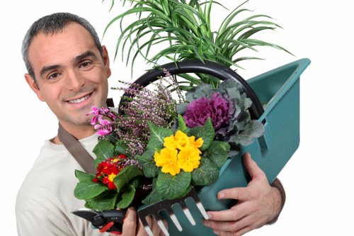 The image shows a professional gardener holding a teal plastic watering can filled with an assortment of colorful flowers, including yellow, purple, and red blooms, alongside lush green foliage. Behind the flowers, there is a vibrant, healthy green potted plant with long, narrow leaves, indicating attentive plant care. The gardener is smiling warmly and dressed in a neutral-colored shirt, suggesting a friendly and professional approach to garden maintenance. The background appears to be an outdoor garden space with natural lighting, possibly on a sunny day, highlighting the lush, well-maintained garden environment typical of professional landscaping and gardening services in Marylebone. The scene emphasizes the focus on outdoor plant care and garden aesthetics, aligning with the services provided by Landscapers Marylebone, and subtly referencing the local area in central London known for its stylish garden designs and outdoor spaces.