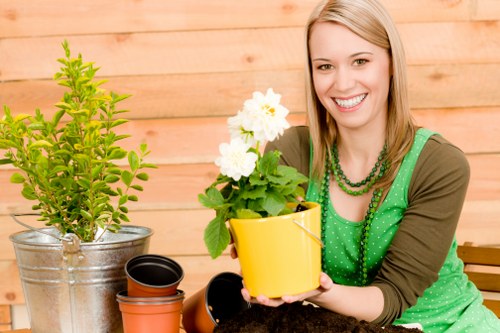 A woman with blonde hair, wearing a green polka-dot apron and a brown long-sleeved shirt, is smiling while holding a yellow flowerpot with white flowering plants in a garden setting. The background features a light wooden fence, with a variety of gardening elements visible, including a large metal watering can with a lush green shrub, an orange plastic pot, and a smaller black plant pot. The outdoor space appears well-maintained, with a focus on healthy plant growth and a tidy layout. The scene is illuminated by natural daylight, suggesting a bright, clear day, and the overall environment reflects a suburban gardening scene typical of Marylebone or nearby London areas, supporting gardening and landscaping services offered by landscapersmarylebone.co.uk. The arrangement emphasizes a welcoming, vibrant outdoor space suitable for gardening activities aimed at enhancing urban gardens and outdoor aesthetics.