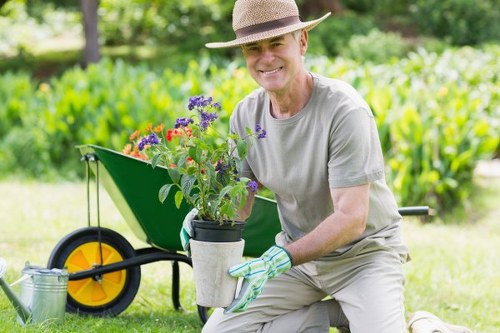 A middle-aged man dressed in a light grey T-shirt, beige trousers, and a straw hat is kneeling on a well-maintained grassy lawn in a garden, smiling at the camera while holding a potted flowering plant with purple, red, and green foliage. In the background, there is a lush green hedge and a wheelbarrow with a yellow wheel and black handles, filled with gardening tools and supplies, situated on the neatly trimmed lawn. The garden features a mix of vibrant plants, with a mix of soil beds and paved pathways, creating a tidy outdoor space suitable for gardening and landscaping maintenance. The scene captures a bright, clear day, highlighting the natural tones of the garden and the outdoor environment. This setting exemplifies professional gardening and landscaping services offered by Landscapers Marylebone, emphasizing plant care, garden planting, and outdoor beautification within the local area near postcode W1.