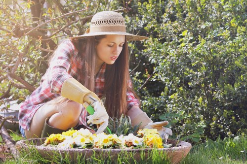 A woman wearing a wide-brimmed straw hat, a plaid shirt, and gardening gloves is kneeling on a grassy lawn in a garden, tending to a colorful flower bed with various yellow, white, and purple flowers. The garden features a neatly maintained lawn with dense green foliage and trees in the background, providing a lush outdoor environment. The scene is illuminated by natural daylight, highlighting the natural tones of the plants and the vibrant colors of the flowers, suggesting a clear, sunny day suitable for gardening activities. This setting, part of a landscaped outdoor space, exemplifies typical garden elements such as flower beds and grass, and aligns with gardening services offered by companies like Landscapers Marylebone, supporting local outdoor maintenance and landscaping in London postcodes including NW1, within the Marylebone area.