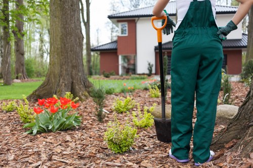 The image depicts a landscaped garden area in Marylebone with a mature tree in the foreground, surrounded by a neatly mulched garden bed containing vibrant red tulips and small green shrubs. In the background, there is a well-maintained lawn bordered by additional trees and a modern residential building with red brick and large windows. A person dressed in green gardening overalls is partially visible, holding a garden tool, likely engaged in planting or maintenance work. The scene is set outdoors on a cloudy day, providing soft, natural light that highlights the greenery and natural tones of the garden elements. The outdoor space appears to be a back garden or landscaped yard designed for outdoor enjoyment and gardening activities, showcasing areas suitable for lawn care, planting, and garden maintenance—services offered by Landscapers Marylebone. The overall environment is tidy and inviting, emphasizing professional landscape care that enhances the natural beauty of the outdoor space in Marylebone.