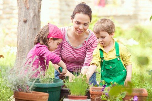 A woman with dark hair tied back, wearing a pink and white striped long-sleeve top, is gardening outdoors in a residential garden with two young children. The girl, dressed in a pink jacket and headscarf, is planting or tending to plants in a green pot, while a boy wearing a yellow shirt and green overalls watches and participates. The garden features a variety of potted plants and flower beds with blooming flowers and lush foliage. In the background, there is a large tree trunk, soft natural light, and a blurred garden scene with greenery and a brick boundary wall, indicating a well-maintained outdoor space suitable for gardening activities. The environment appears calm and inviting, with emphasis on outdoor recreational and gardening activities, reflecting professional gardening expertise such as that provided by Landscapers Marylebone, especially relevant to garden maintenance in London suburbs or similar areas.