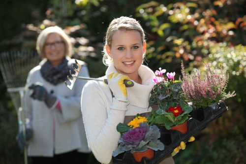 A young woman with blonde hair, wearing a white sweater and yellow gardening gloves, is holding a black tray filled with various colorful flowering plants and greenery in a landscaped outdoor garden. The garden features a well-maintained lawn with dense, vibrant green grass in the foreground, bordered by a flower bed with blooming pink, purple, and red flowers, alongside shrubbery and small ornamental plants. Behind her, an older woman with glasses, grey hair, and a patterned scarf around her neck is slightly out of focus, holding gardening tools and smiling. The background displays a mix of mature trees and bushes with foliage in seasonal shades, illuminated by natural sunlight on a clear day, creating a bright and inviting outdoor scene. This setting highlights professional gardening and planting services, similar to those offered by Landscapers Marylebone, suitable for enhancing residential gardens in the Marylebone area or nearby London suburbs, with a focus on vibrant plant displays and well-maintained outdoor spaces.