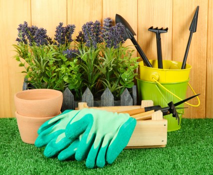 An organized gardening tools and supplies display set up on a green artificial turf surface, featuring a row of potted lavender plants with purple flower spikes and green foliage, two terracotta plant pots stacked together, a light wooden storage box containing a pair of bright turquoise gardening gloves and a small hand rake, a yellow plastic bucket holding black-handled garden tools including a trowel and pruners, all against a background of vertical wooden fence panels typical of a Marylebone garden or outdoor space. The scene suggests preparation for gardening or maintenance activities in an outdoor garden environment, with natural lighting indicating a clear day and the arrangement highlighting a neat, functional outdoor gardening area serviced by local experts like Landscapers Marylebone.